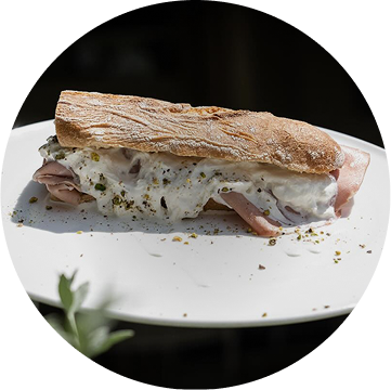 Servizio panino al banco presentato con una fotografia di un panino fatto con pane toscano, mortadella, burrata e granella di pistacchio, preparato dal ristorante da Alcide a Venturina, in Toscana.