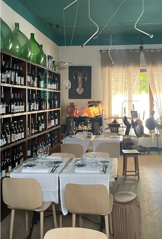 Dettaglio sala interna del ristornate, con tavoli apparecchiati e dispensa di vari vini nel ristorante da Alcide a Venturina, in Toscana. Il ristorante usa Ingredienti toscani e costruisce una storia familiare.